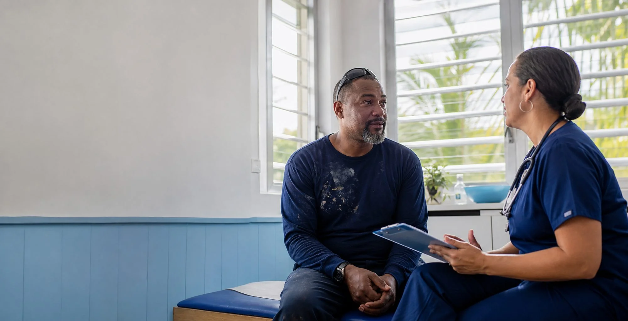 A man in work clothes sits on an exam table talking with a nurse who holds a clipboard, in a bright island clinic room.